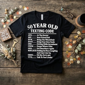 May include: Black t-shirt with the text "50 YEAR OLD TEXTING CODE" and a list of acronyms with their meanings. The shirt is on a wooden surface with scattered confetti and decorative items.