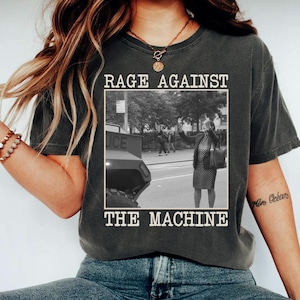 May include: Dark gray t-shirt with the text "RAGE AGAINST THE MACHINE" above and below a black and white image of a person on a street. The shirt is a crew neck style.