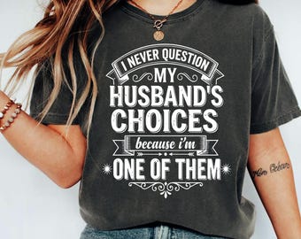 May include: Dark gray t-shirt with the text "I NEVER QUESTION MY HUSBAND'S CHOICES because I'm ONE OF THEM" in white. The shirt has a crew neck and short sleeves. The shirt is worn by a person.