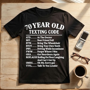 May include: Black t-shirt with white text that reads "70 YEAR OLD TEXTING CODE" and a list of acronyms with their meanings. A flip phone, eyeglasses, and a coffee cup are on the wooden surface.