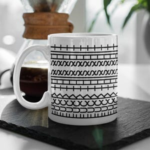 May include: White ceramic coffee mug with a black geometric pattern. The design features horizontal bands of lines, crosses, and wave-like shapes. The mug has a large, curved handle and sits on a dark surface. A coffee maker is visible in the background.