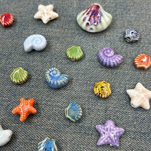 May include: A collection of colorful ceramic seashell and starfish beads in various shapes and sizes. The beads are in shades of red, blue, green, yellow, orange, and purple. The beads are scattered on a denim background.