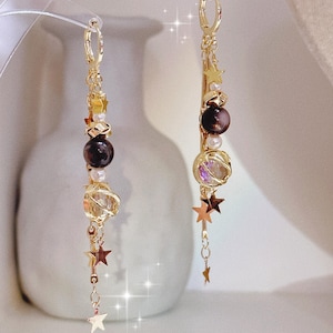 Op de afbeelding: Een paar goudkleurige oorhangers. Elke oorbel heeft donkerbruine bollen, kleine parels en heldere, facetgeslepen stenen. Gouden sterbedels hangen aan de onderkant, wat een hemelse touch aan de sieraden geeft.