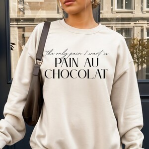 Peut inclure: Sweat-shirt crème avec l'inscription noire "The only pain I want is PAIN AU CHOCOLAT". Il est porté avec une jupe plissée et des bottes blanches. Un sac à bandoulière marron et une tasse à café blanche sont également visibles.