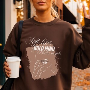 Peut inclure: Un sweat-shirt marron avec le texte "Soft lips, Bold Mind, Mocha inside" et un dessin de lèvres. Le sweat-shirt est porté avec une jupe plissée beige. Une personne tient une tasse à café blanche.