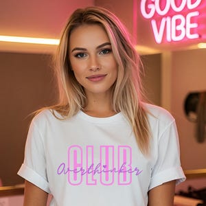 Peut inclure: Un t-shirt blanc avec le texte "CLUB Overthinker" en rose et violet. Le t-shirt est porté par une personne aux cheveux blonds. Une enseigne au néon avec l'inscription "GOOD VIBE" est en arrière-plan.