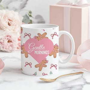 Peut inclure: Mug en céramique blanche avec un cœur rose portant l'inscription « Gentle Mornings ». La tasse est décorée de bonhommes en pain d'épices et de nœuds roses. Une cuillère dorée repose sur une surface en marbre, avec une boîte cadeau rose et des fleurs en arrière-plan.