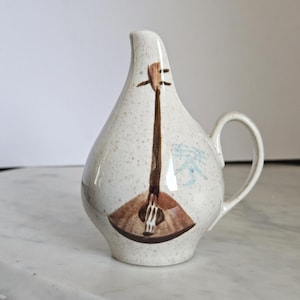 May include: A speckled, off-white ceramic pitcher with a brown and tan lute design on the front. The pitcher has a curved handle and a slender spout. A blue and white floral design is on the side.
