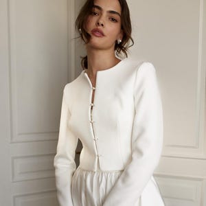 May include: A white bridal ensemble featuring a long-sleeved jacket with pearl-like button closures and a matching skirt. A small, textured white handbag is held in the hands. The outfit is elegant and suitable for a wedding.