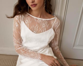 May include: A white dress featuring a sleeveless bodice with a scoop neckline and a sheer, long-sleeved overlay made of intricate floral lace. The lace overlay covers the shoulders and arms, while the solid white fabric forms the base layer.
