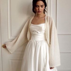 May include: A white satin wedding dress with a square neckline and a full skirt. A cream-colored, open-front bolero-style jacket with pearl trim is worn over the dress. The jacket has long sleeves and is adorned with small pearls.