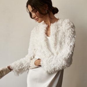 May include: A white, fluffy jacket with a V-neck and long sleeves. The jacket has a textured, shaggy appearance. The model is wearing a white dress and gloves. The overall aesthetic is elegant and sophisticated.
