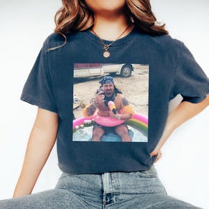 May include: Dark blue t-shirt with a square image of a man in a small pool, wearing a bandana, holding a glass of wine, and sitting in a pink flamingo float. The pool has a rainbow design.