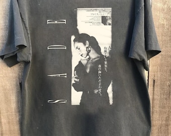 May include: Dark gray t-shirt featuring a black and white image of a woman with large hoop earrings. The shirt has the word "Sade" in white block letters and Japanese text.
