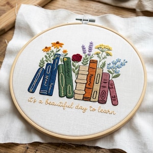 May include: Embroidered hoop art featuring a stack of colorful books with floral accents. The books are in shades of blue, green, orange, and burgundy. The text "it's a beautiful day to learn" is embroidered in gold thread.