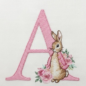 Puede incluir: Letra "A" rosa bordada con un conejito marrón que lleva una chaqueta rosa y rodeado de rosas rosas y hojas verdes. El diseño está sobre un fondo de tela blanca.