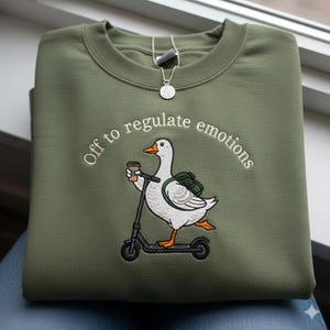 May include: An olive green sweatshirt with the embroidered phrase "Off to regulate emotions." A white goose with a backpack and coffee cup rides a black scooter. A silver necklace with a pendant is also visible.