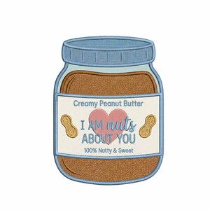 Puede incluir: Un tarro bordado de crema de cacahuete con tapa azul y una etiqueta que dice "I AM nuts ABOUT YOU". La etiqueta también incluye un corazón rosa y dos ilustraciones de cacahuetes. El texto "100% Nutty & Sweet" también está en la etiqueta.