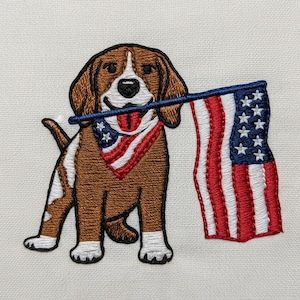 May include: Embroidered image of a brown and white dog wearing a patriotic bandana and holding a US flag in its mouth. The flag features red and white stripes and blue stars. The dog is sitting on a white background.