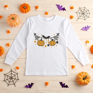 May include: White long-sleeve shirt featuring a Halloween-themed design. The graphic includes two orange pumpkins, bats, spiderwebs, and stars. The shirt is surrounded by pumpkins, candy corn, and decorative bats and spiderwebs.