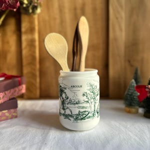 Peut inclure: Un pot à ustensiles de cuisine en céramique blanche avec une illustration verte d'un paysage et le mot "ARCOLE". Il contient des cuillères et spatules en bois. L'arrière-plan comprend une surface en bois et des objets décoratifs.