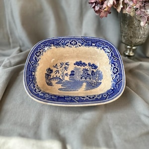 Könnte beinhalten: Ein rechteckiger, antiker Teller aus blau-weißer Keramik. Der Teller zeigt eine detaillierte Landschaftsszene in der Mitte und einen dekorativen Rand. Die Farbpalette ist hauptsächlich blau und cremefarben, mit einer Vintage-Ästhetik. Der Teller dient wahrscheinlich zum Servieren oder Ausstellen.