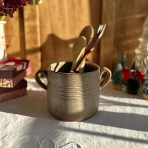 Peut inclure: Une tasse en céramique tachetée de couleur beige avec deux anses, contenant des cuillères et des fourchettes en bois. La tasse présente des lignes gravées horizontales. L'arrière-plan comprend une surface en bois, des fleurs et de petits arbres de Noël.