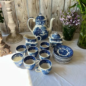 Peut inclure: Un service à thé en porcelaine bleu et blanc, comprenant une théière, une crémière, un sucrier, des tasses et des soucoupes. L'ensemble présente un motif de paysage. Les articles sont disposés sur une nappe blanche, avec un bougeoir et des fleurs en arrière-plan.