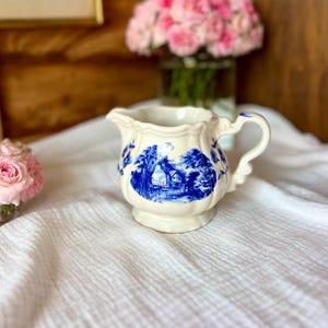 Peut inclure: Crémier en céramique crème avec un motif de paysage bleu. Le crémier a une anse et un bec verseur, et la scène représente une maison et des arbres. Des fleurs roses sont en arrière-plan.