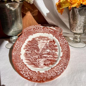 Peut inclure: Assiettes décoratives rouges et blanches avec une bordure florale et une scène champêtre. Les assiettes sont disposées sur une nappe blanche, avec des fleurs en arrière-plan.