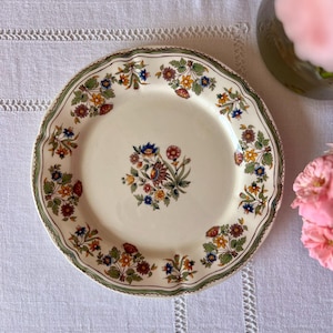 Puede incluir: Un plato de cerámica color crema con borde festoneado y diseño floral. El plato presenta un borde verde con un patrón repetido de flores y follaje coloridos. También es visible un ramo floral central.
