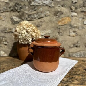 Peut inclure: Un pot en terre cuite à deux anses avec un couvercle assorti, présentant une partie supérieure vernissée brun foncé et une base en argile mate naturelle. Le récipient a un corps arrondi et un petit bouton au centre du couvercle.