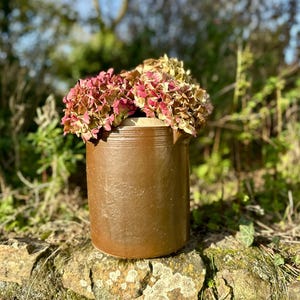 May include: A brown ceramic crock filled with dried hydrangeas. The flowers are a mix of pink and tan hues, contrasting with the crock's earthy tones. The crock sits on a stone wall, with greenery in the background.