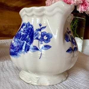 May include: A white ceramic vase with a bulbous body and scalloped rim. The vase is decorated with blue floral and landscape designs. The vase sits on a small, scalloped base. Pink roses are visible in the background.