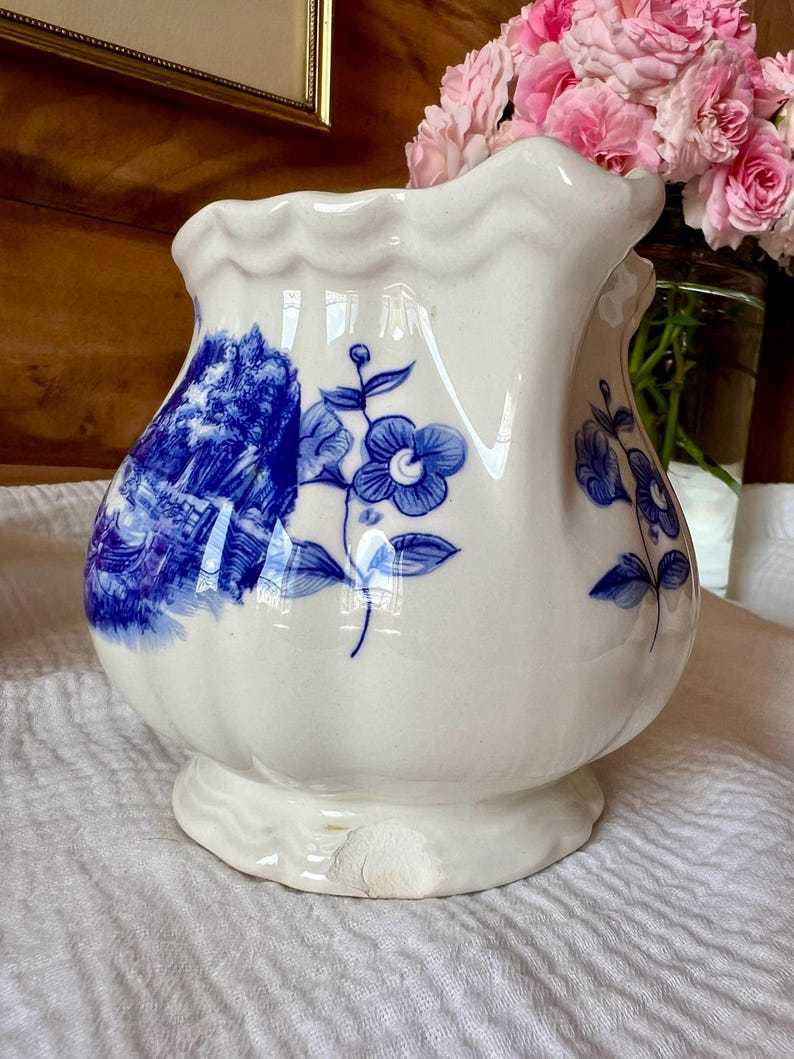 May include: A white ceramic vase with a bulbous body and scalloped rim. The vase is decorated with blue floral and landscape designs. The vase sits on a small, scalloped base. Pink roses are visible in the background.