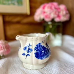 May include: A small, off-white ceramic creamer with blue floral and landscape designs. The creamer has a scalloped rim and a small handle. The creamer is sitting on a white cloth.