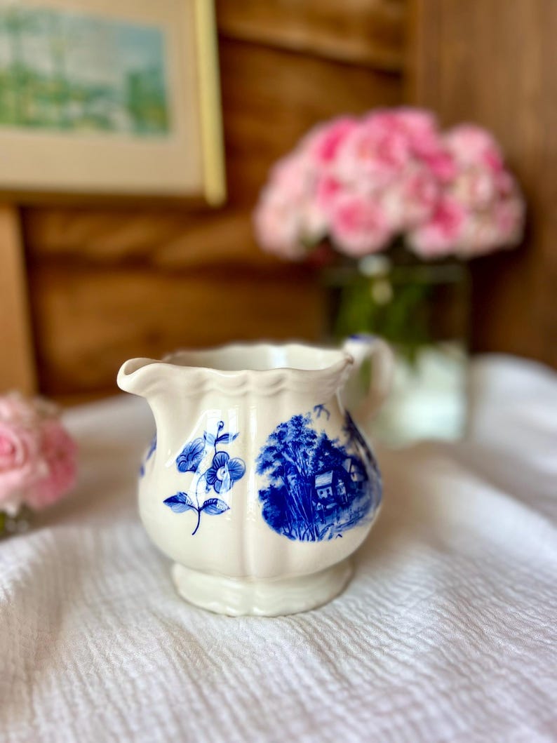 May include: A small, off-white ceramic creamer with blue floral and landscape designs. The creamer has a scalloped rim and a small handle. The creamer is sitting on a white cloth.