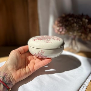 May include: A small, round, cream-colored ceramic trinket box with a lid. The box and lid are decorated with a repeating pattern of red floral designs. The box is held in a hand, resting on a white surface.