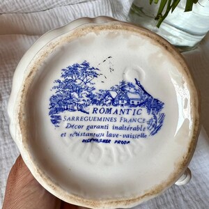 May include: A white ceramic pitcher with a scalloped rim. The bottom features a blue and white transferware design of a rural scene with the text "ROMANTIC SARREGUEMINES FRANCE" and "DISHWASHER PROOF".