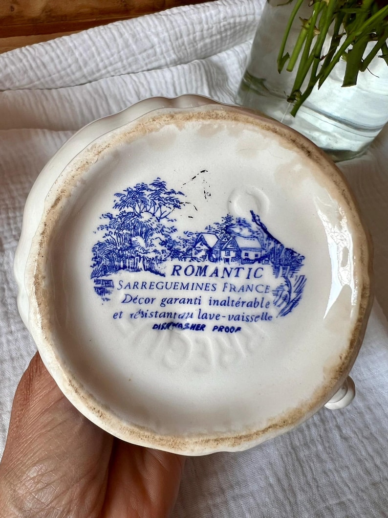 May include: A white ceramic pitcher with a scalloped rim. The bottom features a blue and white transferware design of a rural scene with the text "ROMANTIC SARREGUEMINES FRANCE" and "DISHWASHER PROOF".