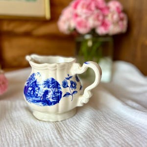 May include: A small, off-white ceramic creamer with a blue and white landscape design. The creamer has a scalloped rim and a decorative handle. A blue floral design is also present. The creamer sits on a white cloth.