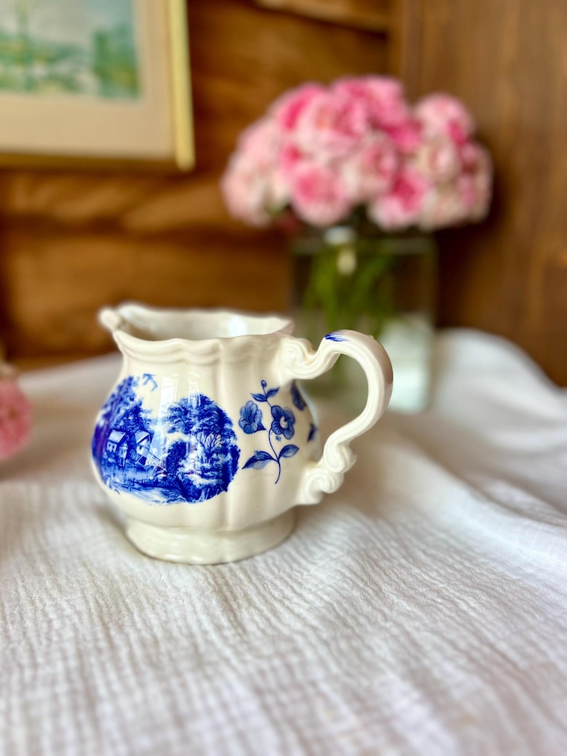 May include: A small, off-white ceramic creamer with a blue and white landscape design. The creamer has a scalloped rim and a decorative handle. A blue floral design is also present. The creamer sits on a white cloth.
