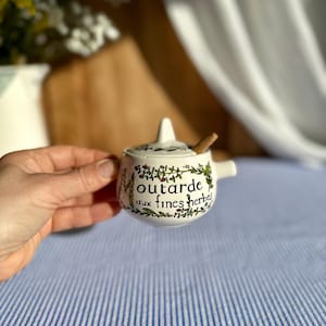 Puede incluir: Un pequeño tarro de mostaza de cerámica blanca con tapa y una pequeña cuchara de madera. El tarro está decorado con detalles florales verdes y rojos y las palabras "Moutarde aux fines herbes".
