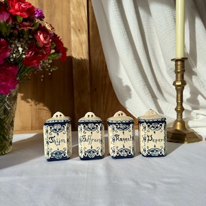 Op de afbeelding: Vier keramische kruidenpotten met deksels, met een crèmekleurige basis en blauwe bloemmotieven. De potten zijn gelabeld met "Thijm", "Saffraan", "Raneel" en "Peper". Een boeket rode bloemen en een messing kandelaar staan op de achtergrond.
