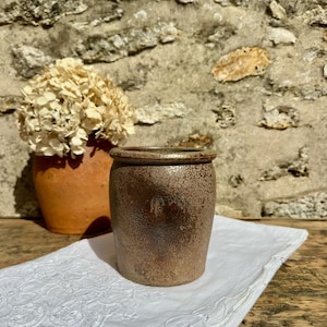 Peut inclure: Un pot en céramique cylindrique avec un vernis moucheté brun et beige, doté d'un bord épais et arrondi ainsi qu'une petite marque estampée discrète sur la face avant. L'objet présente un aspect terreux texturé avec des nuances variées.