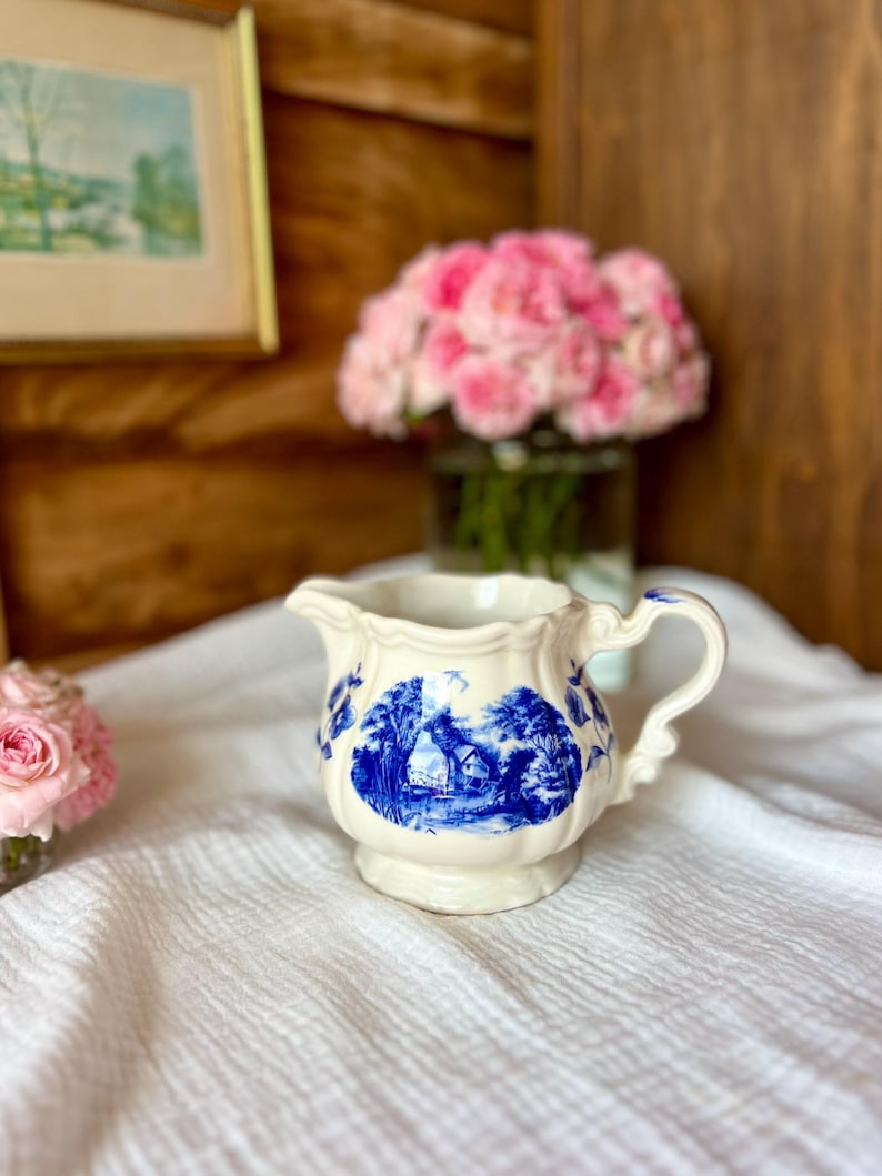 May include: Cream-colored ceramic creamer with a blue landscape design. The creamer has a handle and a spout, and the landscape scene depicts a house and trees. Pink flowers are in the background.