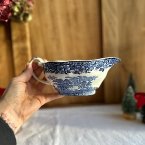 Peut inclure: Un saucier en céramique blanc et bleu avec une anse et un bec verseur. Le saucier présente un motif floral et paysager bleu sur le bord et sur le corps. Le saucier est tenu dans une main.