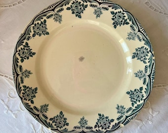 May include: Cream-colored ceramic plate with a teal floral pattern around the rim. The plate features a scalloped edge and a central design of teal roses. The plate is set on a white, embroidered tablecloth.