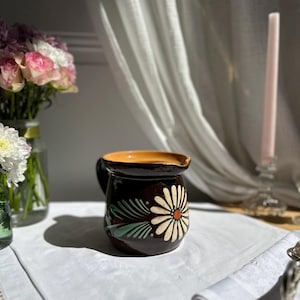 Pode incluir: Um jarro de cerâmica castanho com um desenho floral. O jarro apresenta uma margarida branca com um centro laranja e folhas verdes. A borda interior é castanha clara. O jarro está sobre uma toalha de mesa branca.
