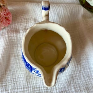May include: Cream-colored ceramic creamer with blue floral patterns and a spout. The handle is accented with blue paint. The creamer is empty and sits on a white, textured surface.
